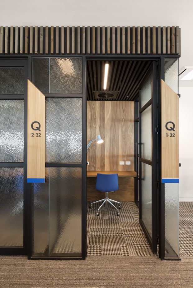 A compact enclosed office pod or meeting booth within a commercial interior. The pod has black metal framing, textured translucent glass panels, light timber doors with signage, and a dark slatted ceiling. Inside is a small timber desk, blue office chair, task lamp, patterned carpet, and wood-panelled rear wall. The surrounding office uses neutral carpet and bright white finishes.