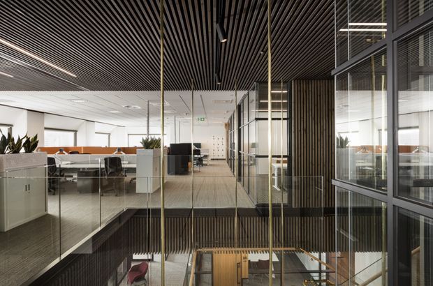 A contemporary commercial office interior viewed across a double-height atrium or stair void. Open-plan workstations with desks, chairs and white storage partitions occupy the left side, while extensive black-framed glazing and dark vertical timber slatted wall and ceiling elements define the right and upper areas. A stair or lower-level circulation zone is visible below, with bright linear lighting and a neutral carpeted floor creating a refined, professional workspace.