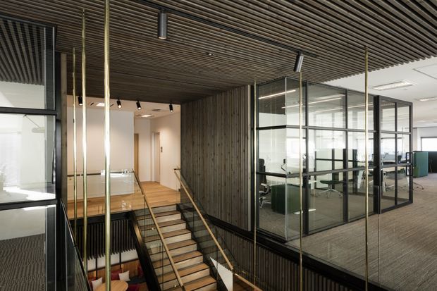 Contemporary commercial office interior with a dramatic stairwell, mezzanine-level workspaces enclosed in black-framed glass partitions, and a timber batten ceiling and feature wall. Brass-toned slender columns and handrails contrast with the dark timber and glazing, while open-plan desks are visible beyond the meeting rooms.