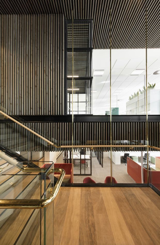 A multi-level commercial interior viewed from a stair landing or mezzanine. The space features warm timber flooring and extensive vertical timber slat wall and ceiling lining, black metal framing and balustrades, clear glass panels, and brass-toned handrails. An open stair and atrium connect to a bright office area below, with red upholstered seating and workspaces visible. The design has a refined contemporary character with strong linear detailing and a double-height void.