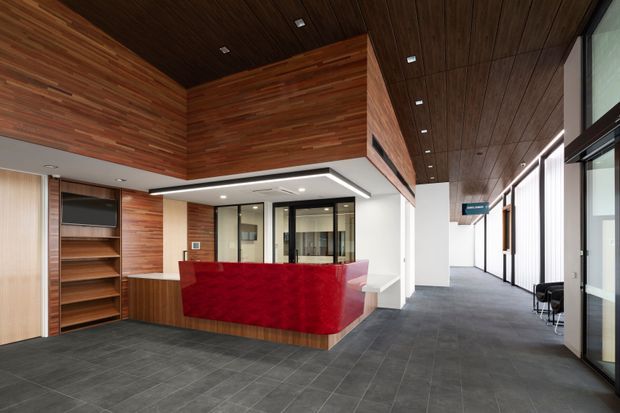 A contemporary commercial reception and lobby interior with a curved red reception desk, dark grey tiled flooring, extensive warm timber wall and ceiling lining, built-in timber shelving with a display screen, and glazed office or corridor partitions. A long circulation corridor extends to the right with seating and full-height glazing. The space is clean, refined, and moderately bright, with strong horizontal timber detailing and integrated recessed lighting.