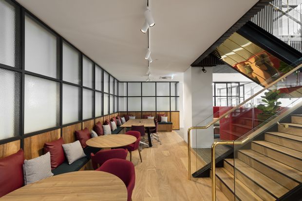 A contemporary commercial interior with a café or hospitality seating area beside a prominent open stairway. The room features curved light-wood tables, burgundy upholstered chairs, a long built-in bench with colourful cushions, timber flooring and wall panelling, black-framed translucent glass partitions, and exposed stair construction with a brass-toned handrail. Track lighting and a glazed upper-level void add to the modern architectural character.