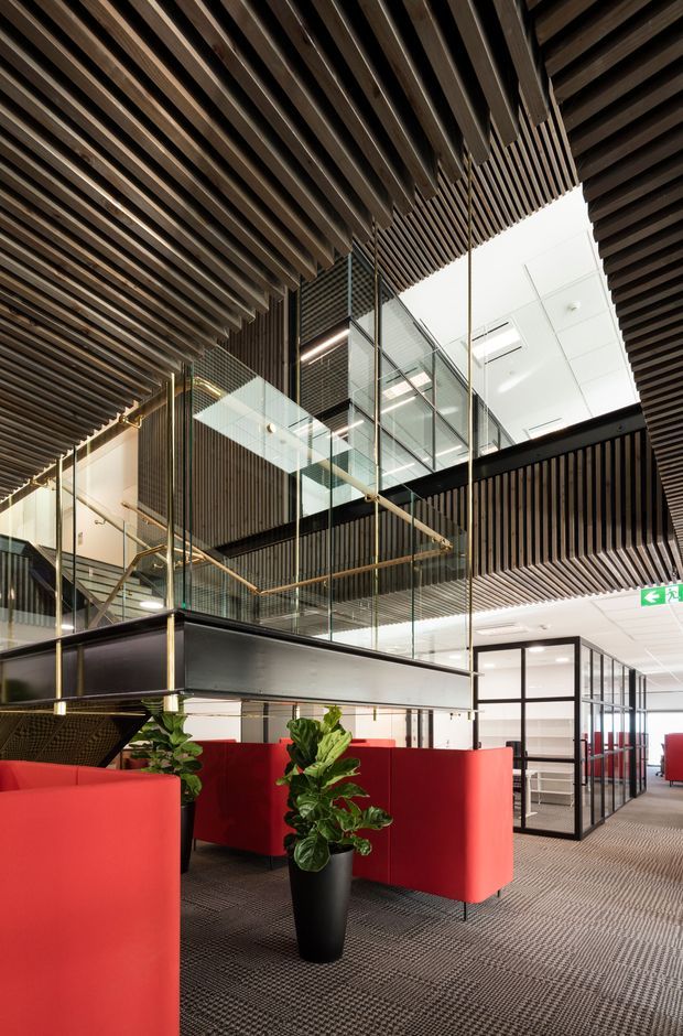 A contemporary multi-level commercial office interior with red upholstered lounge partitions, black-framed glazed meeting rooms, dark timber slatted ceilings and wall screens, glass balustrades, and exposed internal voids. Brass-toned railings and indoor plants add warmth to the otherwise dark, neutral fit-out. The image emphasizes the open atrium and layered office circulation spaces.
