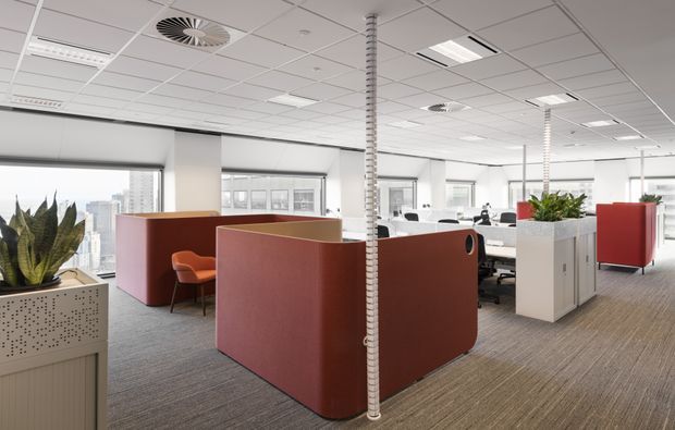 Bright open-plan commercial office interior with carpeted flooring, a suspended acoustic tile ceiling, large windows with roller blinds, white desks and workstations, and several curved rust-red upholstered privacy booths. White planter/storage partitions with indoor plants divide the workspace, while a cityscape is visible through the glazing. The overall environment is contemporary, functional, and softly colourful.
