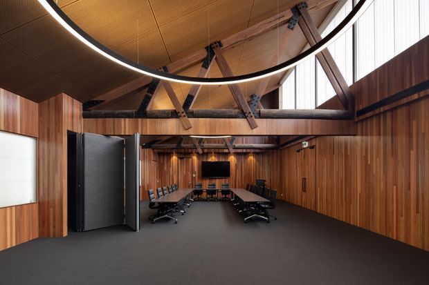 A large contemporary meeting or boardroom with a long conference table, black office chairs, wall-mounted display, dark carpet, and extensive warm timber wall and ceiling lining. Exposed timber structural beams and black steel connector plates span the room, while high clerestory glazing brings in daylight. A large circular suspended LED light forms a prominent overhead feature.