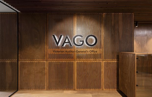A warmly lit commercial interior featuring a large perforated timber wall with framed panels and illuminated VAGO signage reading “Victorian Auditor-General’s Office.” A slatted timber ceiling, timber flooring, and dark timber cabinetry reinforce the sophisticated wood-clad fit-out.