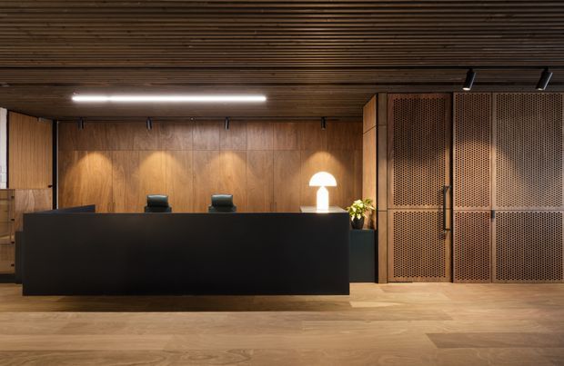 A dark, sophisticated reception desk dominates a warm timber-lined interior. The space features a long matte-black counter, timber wall and ceiling panelling, integrated linear and directional lighting, black task chairs, a small table lamp and plant, and large perforated timber or wood-toned cabinet panels on the right. The composition resembles a refined commercial reception or lobby with restrained, contemporary detailing.