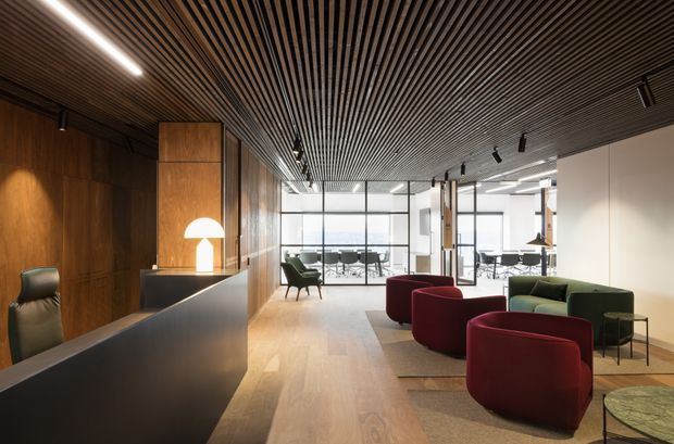 A sophisticated contemporary commercial reception and lounge area with a dark timber reception desk and wall panelling, warm timber flooring, and a striking slatted timber ceiling. Deep burgundy and olive-green lounge chairs sit on a neutral rug, while glazed meeting or dining spaces extend toward the rear. Linear and track lighting create a polished, refined atmosphere.