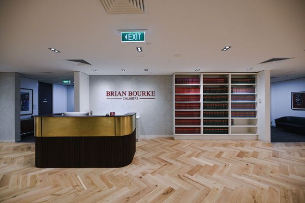 A refined law chambers reception area with a curved dark timber and brass reception desk, herringbone timber flooring, pale walls, recessed lighting, and a large built-in shelving wall filled with law books. The Brian Bourke Chambers logo is displayed on the central wall, with adjoining corridors and seating areas visible to either side.