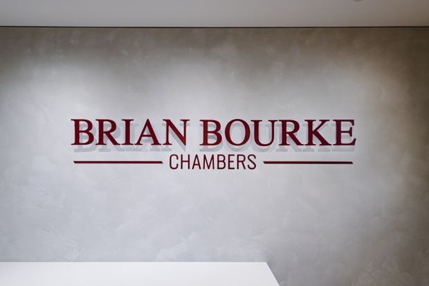 A close-up interior view of a textured light-grey plaster wall featuring the maroon lettering “BRIAN BOURKE CHAMBERS” with horizontal lines above a white reception or counter surface at the bottom edge. The image appears to be signage within a professional commercial interior.