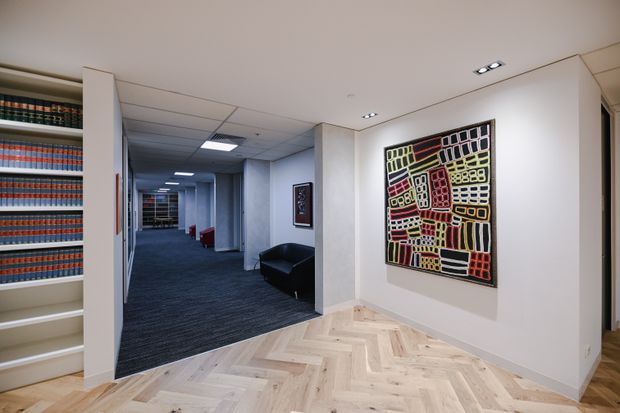 A contemporary institutional or commercial interior showing a light herringbone timber-floored foyer opening onto a carpeted corridor. Built-in shelving with colourful books is visible at left, while a large geometric artwork hangs on a pale wall. Recessed ceiling lighting, white walls, and a black lounge chair create a clean, refined reception or hallway setting.