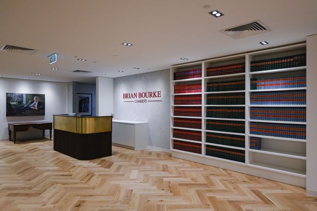 A refined law firm reception area with a large built-in bookshelf filled with colour-coded law books, a curved reception desk with a metallic gold front and dark base, white cabinetry, wall branding, recessed ceiling lighting, and a light timber herringbone floor. The space has a polished, professional commercial interior with restrained contemporary detailing.