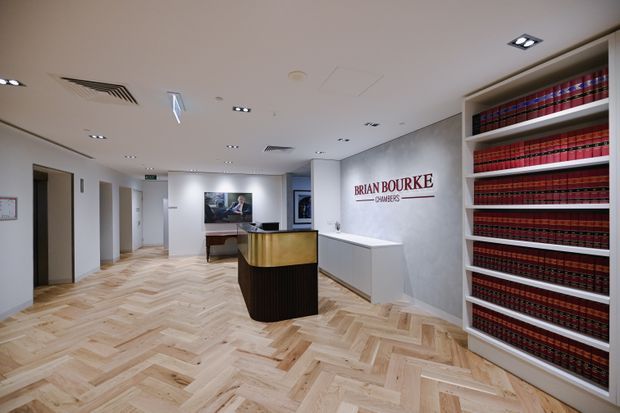 A refined law firm reception area with a curved dark timber reception desk, pale stone or solid-surface counter, white built-in cabinetry, and a large open shelving unit filled with uniformly bound red and black law books. The space has a light timber herringbone floor, white walls and ceiling, recessed downlights, and a grey textured feature wall bearing the Brian Bourke Chambers logo. The overall design is clean, professional, and contemporary.