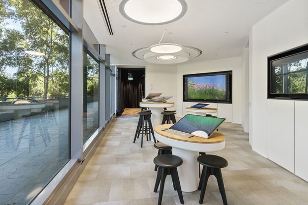 Bright contemporary collaboration or meeting space with floor-to-ceiling glazing, multiple rounded white worktables, black stools, integrated display screens, and distinctive circular ceiling lighting. The room overlooks a landscaped outdoor area and uses a restrained palette of white, pale stone, light timber, and black accents.