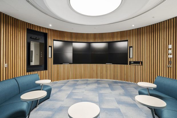 A rounded, acoustically treated media or presentation room with full-height vertical timber slat wall lining, a large central multi-panel display, blue curved built-in seating, small white round tables, pale blue patterned carpet, and a sculptural circular ceiling feature with recessed lighting.