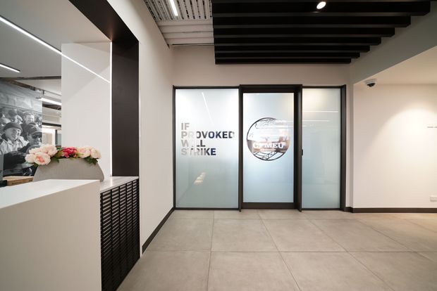 A modern commercial reception or entry lobby with large light-grey tiled flooring, white walls, black-framed frosted glass doors and partitions, and a striking exposed dark timber ceiling battens feature. A white reception counter with floral arrangement and black patterned screen is visible at left, with graphic signage on the glazing and a monochrome wall mural beyond.