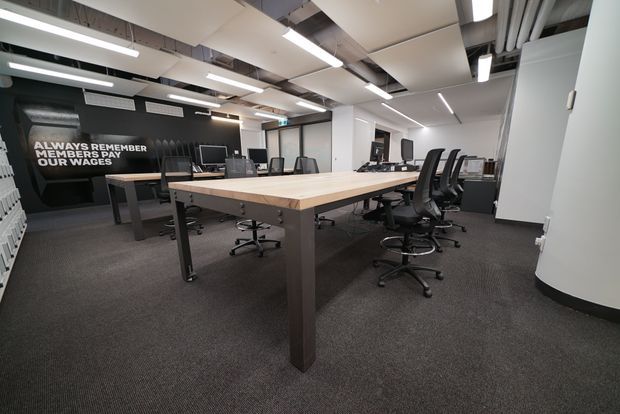 Open-plan contemporary office workspace with a large communal timber-topped meeting or work table, multiple black ergonomic office chairs, computer workstations, dark carpet, black feature walls, and exposed ceiling services. Linear pendant and recessed lighting create a bright, functional commercial interior.