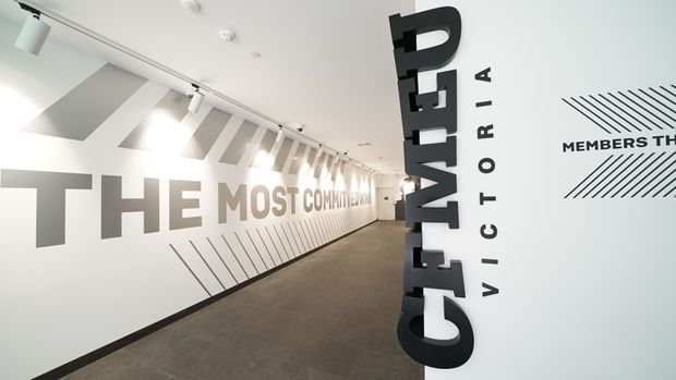 A bright, minimalist commercial corridor or gallery-like foyer with white walls, grey carpet, track lighting, and large black and grey typographic graphics applied to the walls and a prominent vertical column. The space features bold branded signage, diagonal line motifs, and a clean contemporary fit-out.