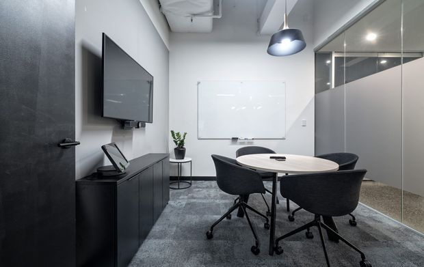 Modern compact meeting room with a round light-wood conference table, four dark upholstered swivel chairs, wall-mounted display, whiteboard, black storage cabinetry, pendant lighting, exposed ceiling services, and glass-partitioned walls. A dark door and textured grey carpet complete the professional office setting.