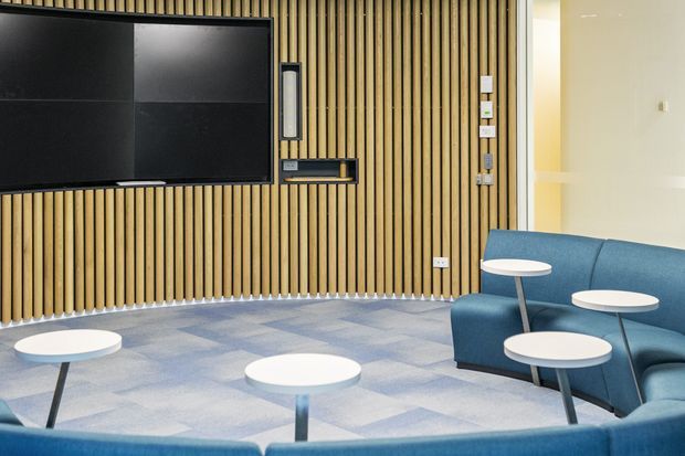 Contemporary commercial lounge or reception area featuring a large wall-mounted display, warm vertical timber slat feature wall, integrated AV and control panels, blue upholstered curved seating, and small round white tables over a patterned blue-grey carpet.