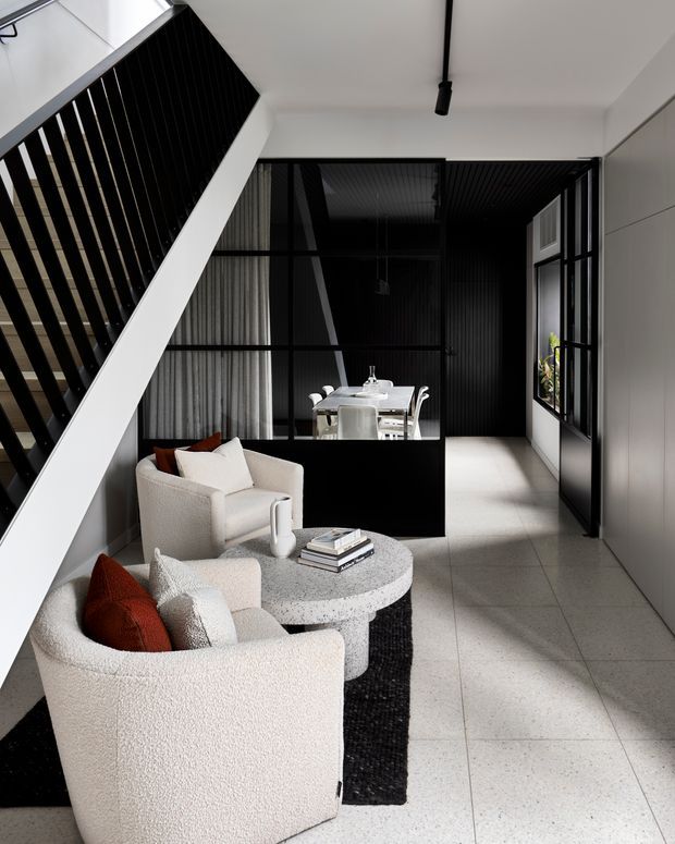 A refined contemporary open-plan interior beneath a staircase, with sculptural cream boucle lounge chairs arranged around a round speckled stone or terrazzo coffee table. Black metal stair balustrading, black-framed internal glazing, dark ribbed wall and ceiling surfaces, and a dining area beyond create strong monochrome contrast. Large pale floor tiles, white walls, sheer curtains, and carefully edited furniture produce a bright, minimalist yet luxurious atmosphere.