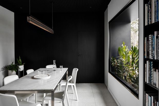 A contemporary dining room with a long pale stone dining table, white chairs, black vertically ribbed wall and ceiling surfaces, a linear pendant light, indoor plants, and a large internal window opening onto greenery. Full-height dark built-in shelving with books occupies the right side.