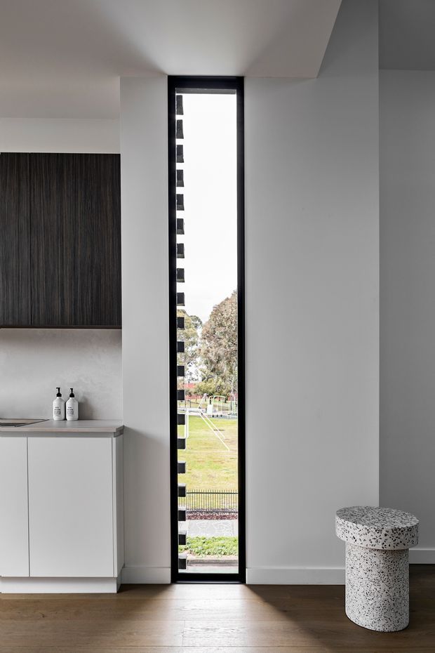 A minimalist interior featuring a tall, narrow black-framed glazed door or window opening onto a landscaped outdoor area. White plaster walls and ceiling contrast with dark vertically textured built-in cabinetry and a simple white vanity or storage unit. The room has warm timber flooring and a small terrazzo cylindrical stool or side table in the foreground.