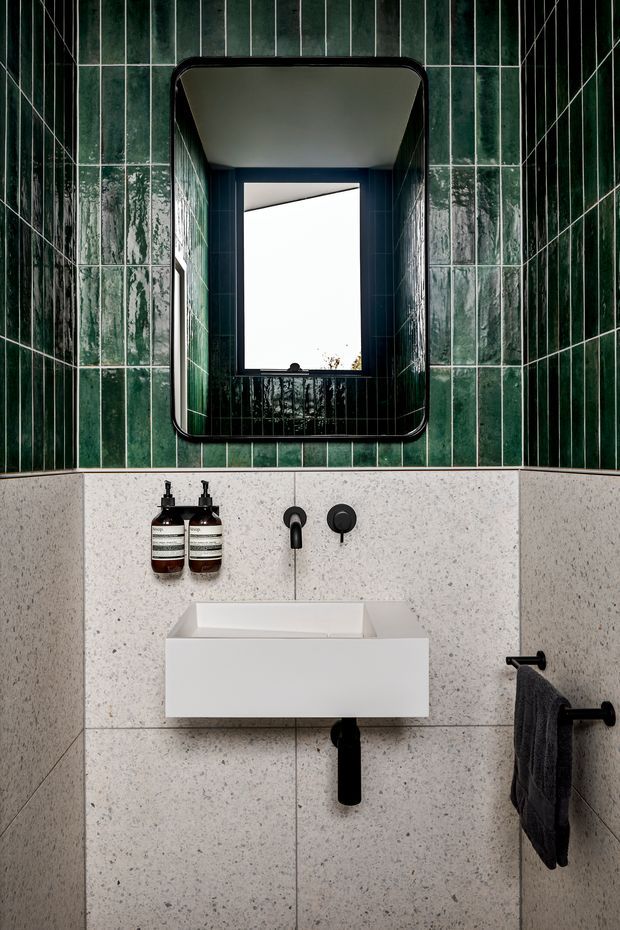 Compact contemporary bathroom with a wall-mounted white basin, black wall-mounted tapware and plumbing, dark towel rail, and amber soap dispensers. The lower walls are clad in pale speckled terrazzo-style slabs, while the upper walls feature glossy vertically oriented deep green tiles with contrasting grout. A rounded rectangular black-framed mirror sits above the basin, reflecting a bright window opening.
