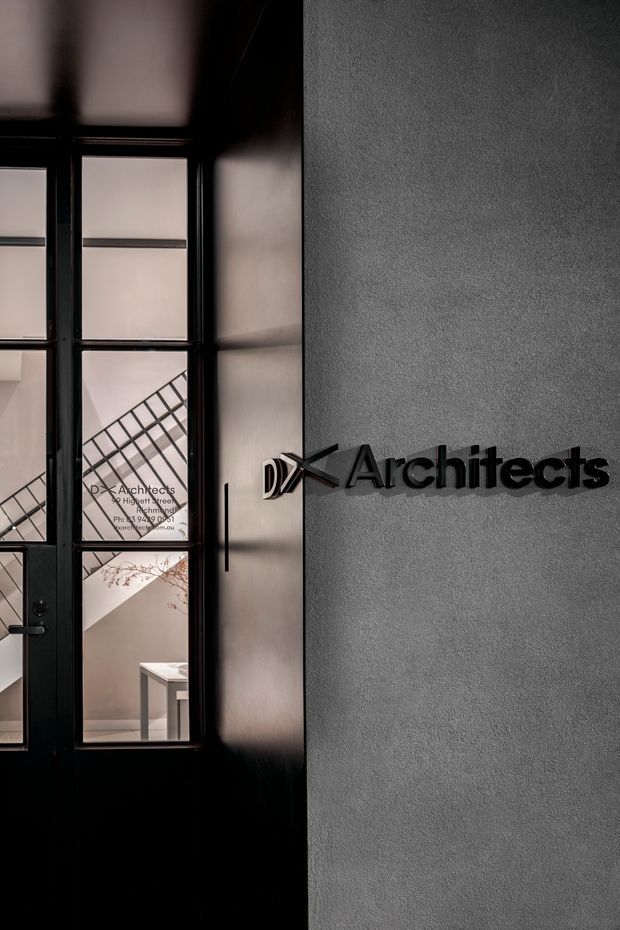 A dramatic close-up of a contemporary architectural office entrance or lobby. A black-framed glazed door and sidelights reveal an interior stair and part of a table, while a textured grey wall carries raised black lettering reading “DX Architects.” The composition is monochromatic, refined, and strongly graphic, with directional light and deep shadows.