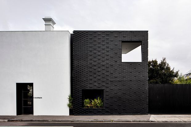 Street-facing urban building facade with a stark white rendered wall adjoining a dark brick-clad rectangular volume. The dark volume has two recessed square openings, while the white section includes a narrow black entrance door and a simple chimney projecting above the roofline. A black vertical-slatted fence continues to the right. The composition is minimalist and contemporary, with a restrained monochrome palette and clean geometric forms.