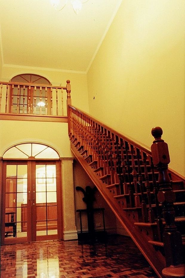 Grand traditional interior entry hall featuring a prominent dark timber staircase with turned balusters, ornate newel posts, and an upper landing. The space has warm pale yellow walls, high ceilings, arched fanlight windows and doors, decorative trim, and a polished parquet timber floor.