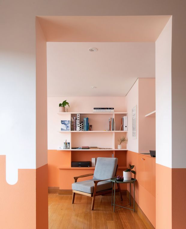 A compact, built-in home study nook framed by a wide architectural opening. The room features pastel pink walls and ceiling, a vivid coral-orange lower wall and fixed cabinetry, open shelving with books and decor, a small desk/work surface, and a mid-century-style upholstered armchair with side tables. Warm timber flooring and soft natural light create a playful yet refined workspace.