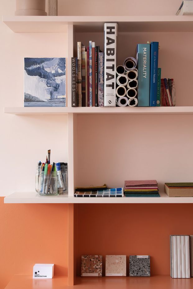 A close-up of a built-in display and storage shelving unit with a warm pink and terracotta colour scheme. The shelves contain architecture and design books, rolled material samples, art supplies, swatches, and stone or terrazzo sample boards. The fixed shelving has clean, simple lines and a matte painted finish, creating a curated design-studio workspace display.