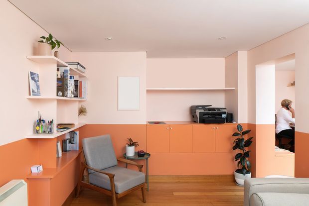 A warm, compact home office with peach-pink walls, a vivid terracotta-orange lower wall and built-in cabinetry, open shelving filled with books and objects, a timber desk, printer, potted plants, a grey upholstered armchair, and timber flooring. An adjoining room is visible through the doorway on the right.