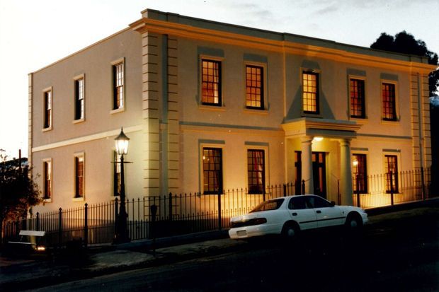 Two-storey rendered masonry building photographed at dusk, with a formal symmetrical facade, tall multi-pane windows, strong horizontal cornice detailing, pilasters, and a columned recessed entrance. Warm interior lighting glows through the windows, while a black metal picket fence and traditional street lantern reinforce the historic classical character.