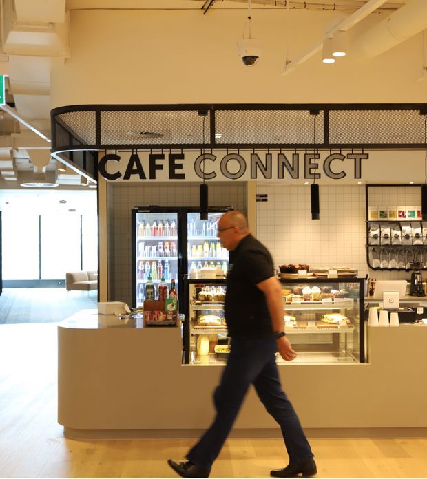 Interior view of a contemporary café counter branded “CAFÉ CONNECT.” The space features a curved light-colored service counter, tiled wall backdrop, refrigerated beverage displays, pastry cases, retail shelving, black metal mesh screening, exposed ducts and services, pendant and recessed lighting, and pale timber flooring. A person is walking across the foreground.