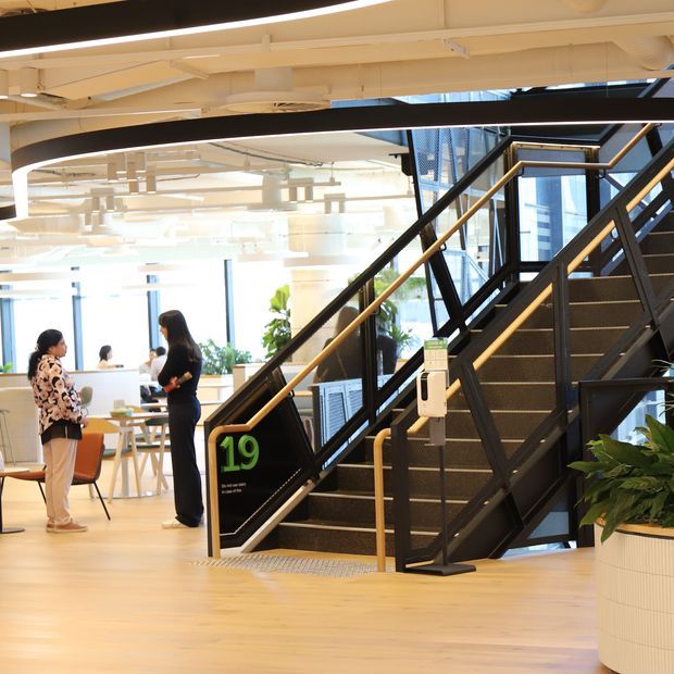 Bright contemporary commercial interior featuring a prominent black metal staircase with timber handrails, light timber flooring, exposed white services and curved black ceiling elements. An open workspace with tables, chairs, people, greenery and large glazed windows is visible behind the stair.