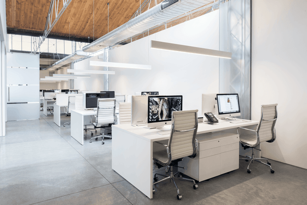 Bright open-plan modern office with white workstations, desktop computers, ergonomic rolling chairs, suspended linear pendant lights, exposed timber ceiling, and exposed metal ductwork. The space has polished concrete flooring and a clean industrial-contemporary fit-out.