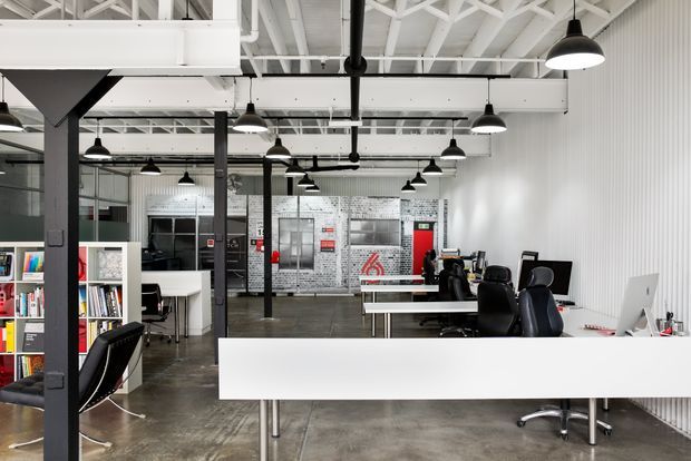 An open-plan creative office with exposed white-painted roof trusses, black structural columns, extensive black pendant lighting, polished concrete flooring, white workstations and desks, and black leather office chairs. Shelving, meeting areas, and graphic wall treatments create an industrial studio atmosphere.