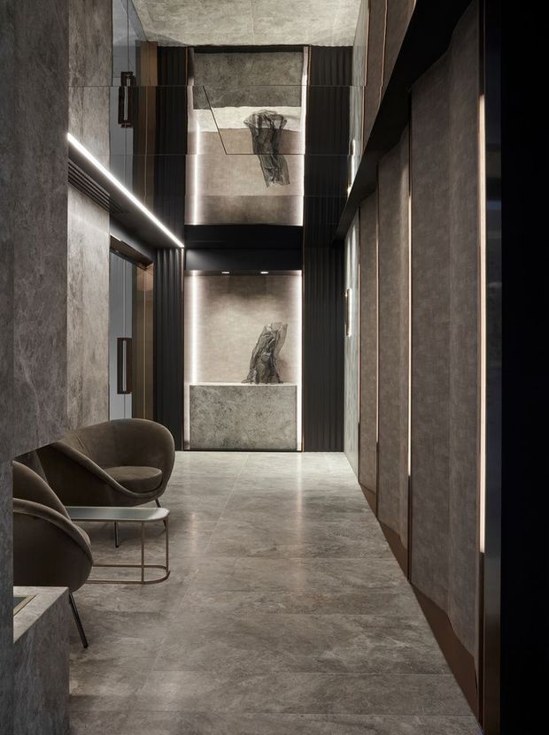A dramatic, narrow interior corridor or lobby finished in dark grey stone-look surfaces. Large-format flooring and wall panels create a continuous monolithic appearance, complemented by black ribbed vertical screens, concealed linear lighting, and dark bronze or black-framed doors. Sculptural artwork is displayed at the far end, while curved upholstered lounge chairs and a slim metal coffee table form a small waiting area along the left side. The space has a refined, moody, gallery-like atmosphere with a tall ceiling or strong vertical volume.