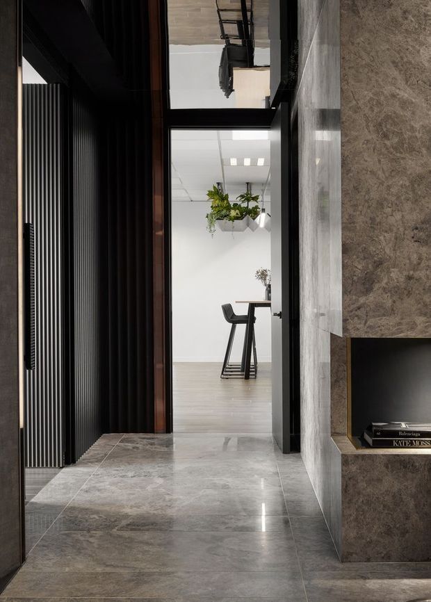 A refined contemporary interior hallway with large-format grey stone flooring and matching stone wall cladding. A dark vertically ribbed feature wall and concealed doorway frame the view into a bright, minimalist workspace with a small table, stools, pendant greenery and a white grid ceiling. A recessed dark stone display niche with books is visible on the right.