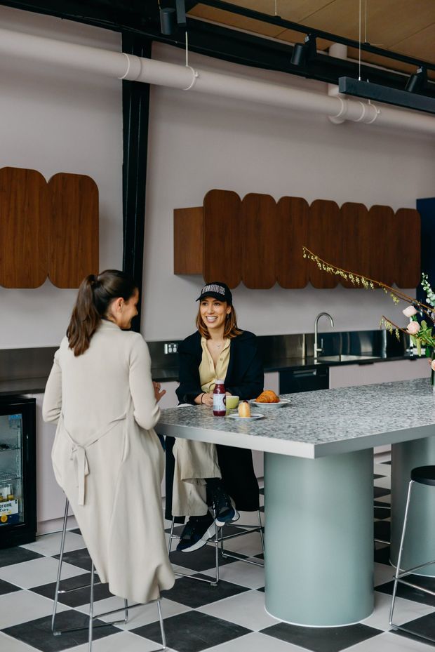 A stylish commercial kitchen or hospitality space with a large speckled stone island, integrated sink and faucet, dark cabinetry, rounded timber wall panels, exposed black ceiling services, track lighting, and a bold black-and-white checkerboard floor. Two people sit and stand at the island, which is furnished with contemporary bar stools and casual food and drink.