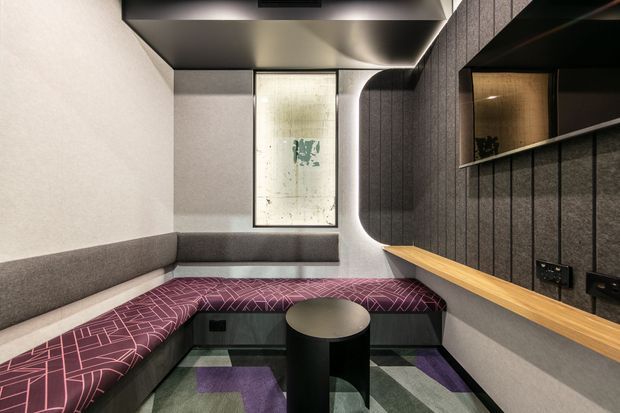 Compact, dark-toned media or lounge room with an L-shaped upholstered banquette, patterned purple seating, small round central table, built-in timber ledge, acoustic wall panelling, integrated shelving, and a large wall-mounted artwork or screen. The room has a contemporary, refined fit-out with concealed and indirect lighting.