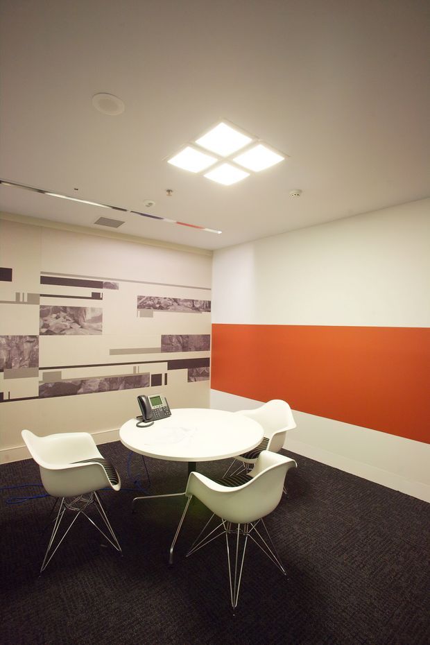 A compact contemporary meeting room with a round white conference table, four white molded chairs, and a desk telephone. The room has dark carpet flooring, off-white walls and ceiling, a bold orange horizontal wall band, and abstract geometric photographic wall graphics. Recessed and geometric ceiling lighting provide bright illumination.