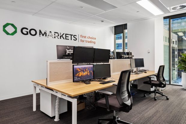A bright modern commercial trading office with multiple computer workstations, monitors, ergonomic office chairs, timber desks, and white built-in cabinetry. The GoMarkets branding is displayed on the wall, with glazed windows providing views of neighbouring office buildings.