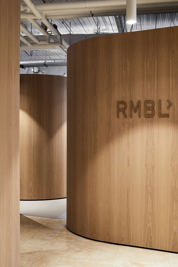 Interior architectural view of a contemporary commercial space with large curved timber-veneered partition walls, integrated directional signage, polished light stone or tile flooring, and an exposed industrial services ceiling with ducts, pipes, and suspended lighting.