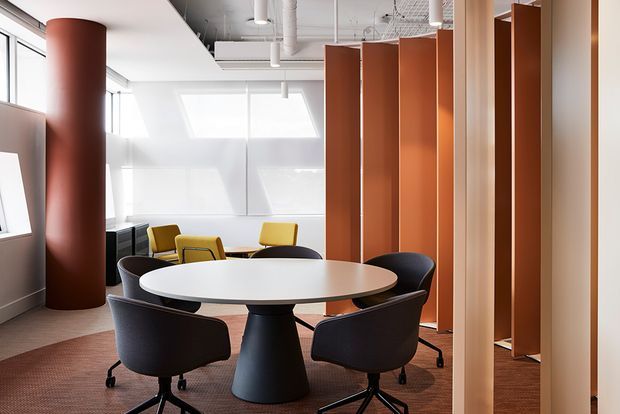 Bright contemporary office breakout and meeting area with a round white table, black swivel chairs, yellow lounge chairs, terracotta cylindrical column, and tall terracotta-orange partition screens. The space has exposed white services, roof windows, and carpet flooring.