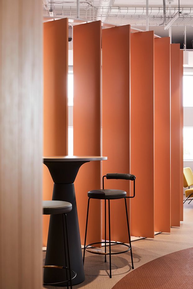A contemporary commercial interior featuring a series of tall terracotta-orange partition panels arranged in a staggered row, creating semi-private zones. Black bar-height stools and sculptural pedestal tables sit on a patterned carpet, with bright glazing and an exposed white services ceiling in the background.