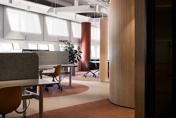 Bright contemporary commercial office interior with open-plan workstations, upholstered privacy screens, task chairs, pendant and linear ceiling lighting, carpeted flooring with circular patterned zones, a curved timber slat wall, and a red cylindrical structural or feature column. Glazed windows and a partially enclosed meeting or workspace are visible in the background.