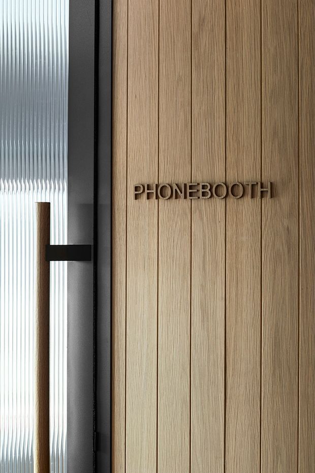 Close-up architectural detail of an office phone booth entrance. The wall is clad in vertically grooved light timber panels with raised dark lettering reading “PHONEBOOTH.” A black metal-framed door with ribbed translucent glass and a dark horizontal handle is visible at left, creating a refined contemporary workplace fit-out.